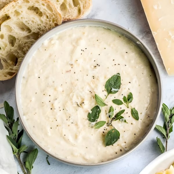 Creamy white dip with herbs and pepper in a bowl, served with sliced bread and Parmesan cheese.