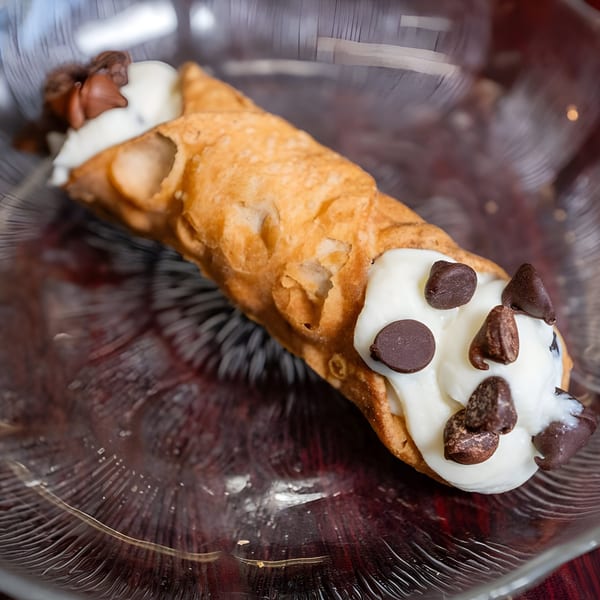 A golden-brown cannoli filled with white cream and topped with chocolate chips in a clear glass dish.